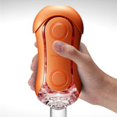 A hand holds a Tenga Flip Orb Sunset Orange by Tenga, a sleek cylindrical pleasure device with a transparent base and circular buttons. The design features clear and textured sections, perfectly blending style with functionality.